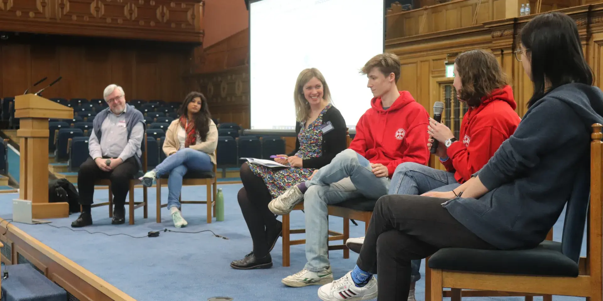 Colour photo of students and staff sat on the stage in the Assembly Hall as they talk to each other as part of a panel discussion